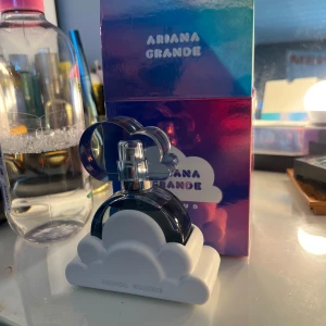 Ariana Grande Cloud Eau de Parfum - Parfymflaska från Ariana Grande i modellen Cloud. Flaskan är rund och genomskinlig med ett blått inslag och har ett unikt lock i form av ett blått moln. Parfymen står i en vit molnformad hållare och kommer i en skimrande blå-lila förpackning. 