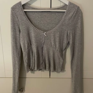 Grå långärmad topp från Gina Tricot - Supersöt grå långärmad topp från Gina Tricot med spetskant vid urringningen och en liten rosett framtill. Modellen är croppad och har en lätt rynkad effekt nedtill. Perfekt att styla med jeans eller kjol!