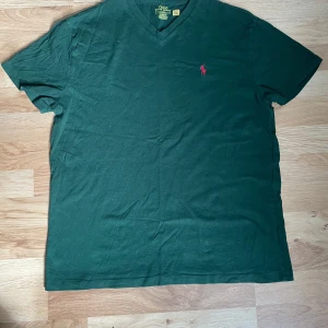 Grön v-ringad t-shirt från Polo Ralph Lauren - Mörkgrön t-shirt från Polo Ralph Lauren med v-ringning och klassisk röd broderad logga på bröstet. Tillverkad i mjuk bomull och har korta ärmar. Perfekt för en avslappnad stil.