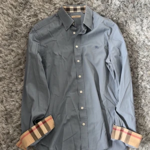 Blå skjorta från Burberry Brit - Snygg blå skjorta från Burberry Brit med klassiska rutiga detaljer på insidan av kragen och ärmsluten. Skjortan har knappar framtill och är långärmad. Perfekt för dig som gillar stilrena plagg med en twist.