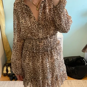 Leopardmönstrad klänning från Zara - Säljer en snygg leopardmönstrad klänning från Zara med långärmad design och volangdetaljer nertill. Klänningen har v-ringning och smockad midja för en bekväm passform. Perfekt för dig som gillar trendiga mönster och vill sticka ut.