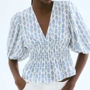 Vit och blå blus med puffärmar och smock - Slutsåld! en vit blus med blått mönster och v-ringning. Blusen har puffiga trekvartsärmar och smockad midja som ger en snygg siluett. Perfekt till både jeans och kjol för en fräsch look.