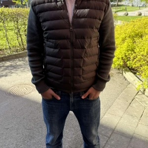 Cardigan - Moncler - Snygg svart dunväst från Moncler med stickade, mörkgrå ärmar och hög krage. Västen har dragkedja framtill och två fickor med dragkedja. Perfekt för lager-på-lager och stilren look.