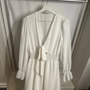 Vit omlottklänning med knyt och smock - Säljer en vit långärmad klänning med omlott och knyt framtill. Klänningen har smock i midjan och volangdetaljer vid ärmsluten. Perfekt för skolavslutningen eller studenten!💞 pris gör och diskuterar💞