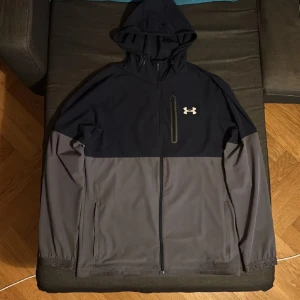 Vindjacka under armour windbreaker - Säljer en snygg hoodiejacka från Under Armour i svart och grått. Jackan har dragkedja framtill, huva och en bröstficka med dragkedja. Perfekt för träning eller chill. Under Armour-logga på bröstet.