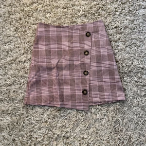 Rutmönstrad Kjol från Gina Tricot - En fin rosa kjol med rutmönster från Gina Tricot i storlek 36, den har knappar längst sidan och en dragkedja där bak för att få på och av sig kjolen, använd endast ett gåtal gånger.