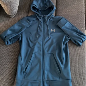 Under Armour tröja - Säljer en blå hoodie från Under Armour med dragkedja och huva. Tröjan har två fickor framtill och en liten logga på bröstet. Perfekt för träning eller chill.