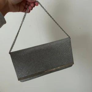 Silver glittrig kuvertväska med kedja - Säljer en snygg silverfärgad kuvertväska med glittrig yta och kedjehandtag. Perfekt att bära i handen eller över axeln. Väskan har en stilren metallist längs kanten för extra detalj.