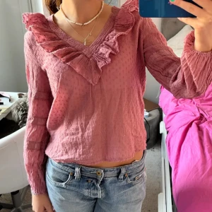 Rosa Neo Noir blus  - Supersöt rosa blus från Neo Noir med v-ringning och volangdetalj längs halsringningen. Långärmad och lätt texturerat tyg som ger en romantisk känsla. Perfekt att styla med jeans eller kjol.