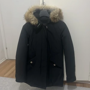 Svart parkajacka med pälskrage från Woolrich - Säljer en svart parkajacka från Woolrich i storlek XS. Jackan har huva med avtagbar pälskant, dragkedja och knappar framtill samt två fickor med lock. Tillverkad i Ramar Cloth (60% bomull, 40% nylon) för extra hållbarhet och skydd mot väder.