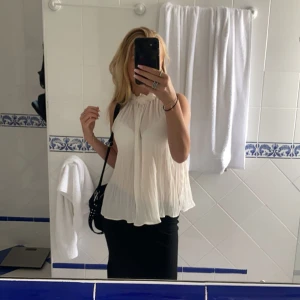 Zara plisserad ärmlös blus - Säljer en luftig och elegant vit blus med plisserat tyg och hög hals. Blusen är ärmlös och har en lös passform, perfekt för varma dagar eller att styla lager på lager.