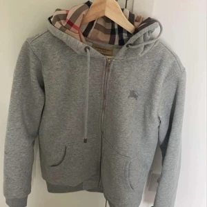 Grå hoodie med dragkedja från Burberry - Säljer en grå hoodie från Burberry med dragkedja och klassiskt rutigt foder i huvan. Tröjan har fickor framtill och justerbar huva med snören. Perfekt för en avslappnad och stilren look nu till kalla sommarkvällar 🤩