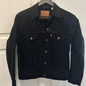 Svart jeansjacka från Levi's - Klassisk svart jeansjacka från Levi's med knappar framtill och två bröstfickor med lock. Jackan har en rak passform och är långärmad. Perfekt att slänga över en hoodie eller t-shirt för en cool look.