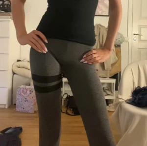STRONGER tränings byxor - Säljer ett par gråa leggings med två svarta horisontella ränder på ena låret. Byxorna har en tight passform och är perfekta för träning eller en avslappnad stil. Mjukt och stretchigt material som sitter skönt hela dagen.