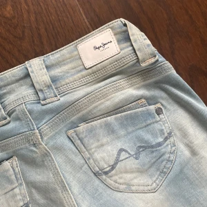  Pepe Jeans - Säljer ett par ljusblå jeans från Pepe Jeans med raka ben och klassisk femficksdesign. Jeansen har dubbla knappar i midjan och diskret broderad detalj på bakfickorna. 