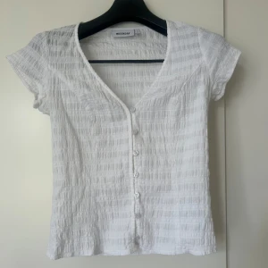 Vit blus med knappar från Weekday - Säljer denna vita topp från Weekday med korta ärmar och v-ringning. Toppen har knappar framtill och modellen är tajt. Perfekt till sommaren! Skriv vid frågor eller funderingar!
