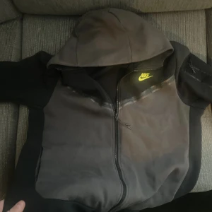 Svart hoodie från Nike med dragkedja - Säljer en svart hoodie från Nike i storlek L. Jackan har huva, hel dragkedja och en gul Nike-logga på bröstet. Perfekt för träning eller chill. Snygg och enkel design med fickor på sidorna.