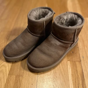Bruna UGG boots i fårskinn - Mysiga bruna UGG boots i klassisk modell med mjukt fårskinnsfoder. Skorna har rund tå, platt sula och är perfekta för kyliga dagar. Utsidan är i mocka och insidan är fodrad med ull för extra värme och komfort.
