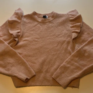 Ljusrosa stickad tröja med volang från H&M - Supersöt ljusrosa stickad tröja från H&M med volangdetaljer på axlarna. Tröjan har långa ärmar och ribbade muddar vid ärmslut och nederkant. Perfekt för att addera lite extra till din outfit!