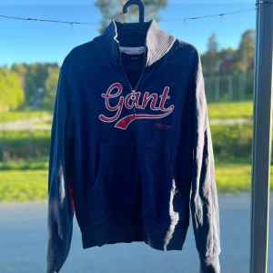 Blå tröja med dragkedja från Gant - Mörkblå tröja från Gant med röd broderad logga framtill och röda detaljer på armbågarna. Tröjan har hög krage, dragkedja och fickor framtill. Perfekt för kyligare dagar. 9/10 i skick perfekt till vår o höst väder