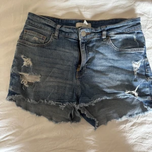 Blå jeansshorts med slitningar - Säljer ett par blå jeansshorts med råa kanter och slitna detaljer framtill. Klassisk femficksmodell med knapp och dragkedja. Perfekta för varma dagar och en avslappnad stil.