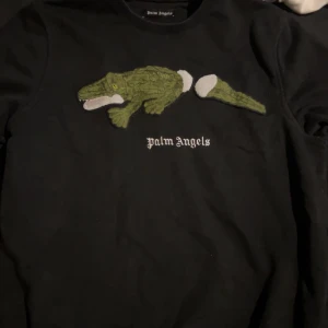 Svart sweatshirt med krokodil från Palm Angels - Säljer en svart sweatshirt från Palm Angels då jag inte längre vill ha den! Den har en grön krokodilapplikation och vit text på bröstet. Tröjan är i 100% bomull och har en klassisk passform med långa ärmar. Skicket är väldigt bra! 