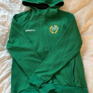 Grön hoodie från Craft med Hammarby IF-emblem - Grön hoodie från Craft med Hammarby IF:s broderade emblem på bröstet. Tröjan har huva, dragkedjefickor och ribbade muddar vid ärmslut och nederkant. Perfekt för fotbollsfans som vill visa sitt stöd.