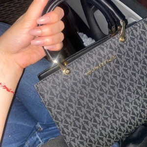 Svart och grå handväska från Michael Kors - Säljer en snygg handväska från Michael Kors i svart och grått med logomönster över hela väskan. Väskan har guldfärgade metalldetaljer och dubbla handtag. Perfekt för dig som vill ha en stilren accessoar. Värde 5000