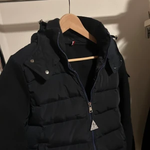 Mörkblå pufferjacka  från Moncler - Säljer en mörkblå pufferjacka från Moncler med avtagbar huva och dragkedja framtill. Jackan har långa ärmar och Moncler-logga på ena ärmen. Perfekt för kyliga dagar och har en sportig look.