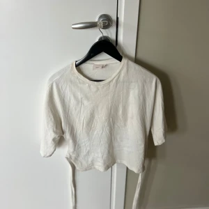 Vit croppad topp med knytband - Säljer en vit croppad topp med knytband vid ryggen. Toppen har lite längre  ärmar och rund halsringning. Perfekt för en avslappnad och trendig stil.