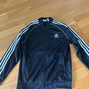 Svart Adidas track jacket med vita ränder - Klassisk svart Adidas track jacket med vita ränder längs ärmarna och broderad logga på bröstet. Jackan har dragkedja framtill, ribbade muddar och två fickor. Perfekt för en sportig och avslappnad stil.