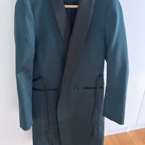 Grönglittrig kavaj med satinrevers från H&M - Snygg grönglittrig  kavaj från H&M med elegant satinrevers och knytband i midjan. Kavajen har en klassisk passform och stängs med en knapp framtill. Perfekt för dig som vill ha en stilren och tidlös look. Kan även användas som klänning 