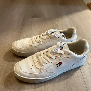Vita sneakers från Tommy Hilfiger - Säljer ett par fräscha vita sneakers från Tommy Hilfiger med klassisk logga på sidan. Skorna har rund tå, platt sula och är tillverkade i syntetmaterial. Perfekta för en clean och stilren look.