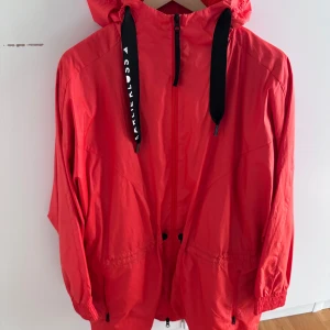 Röd vindjacka från Adidas - Snygg röd vindjacka från Adidas med huva och svarta snören med logga. Jackan har dragkedja framtill, två fickor och resår vid ärmslut och midja. Perfekt för blåsiga dagar. Storlek S. 