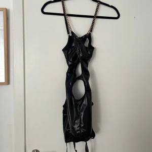 Svart kort klänning med kedjedetaljer - Säljer en svart kort klänning med glansigt material och kedjor som axelband. Klänningen har flera utskurna partier framtill och en tight passform. Perfekt för dig som vill sticka ut med en unik och edgy stil.