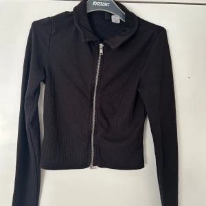 Svart ribbad cardigan jacka med dragkedja - Svart ribbad cardigan jacka med dragkedja framtill och lång ärm. Kortare modell som passar perfekt till vardagslooken. Enkel och stilren design.