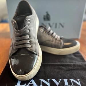 Grå sneakers från Lanvin med lackad tå - Snygga grå sneakers från Lanvin med cool lackad svart tå och vit platt sula. Skorna har snörning och är gjorda i mocka med detaljer i skinn. Perfekt för dig som gillar stilrena och exklusiva sneakers med en unik touch.