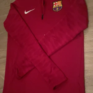 FC Barcelona långärmad träningströja Nike - Röd långärmad fotbollströja från Nike med FC Barcelona-emblem på bröstet och vit Nike-logga. Tröjan har halv dragkedja framtill och är tillverkad i ett lätt och sportigt material som passar perfekt för träning eller match.