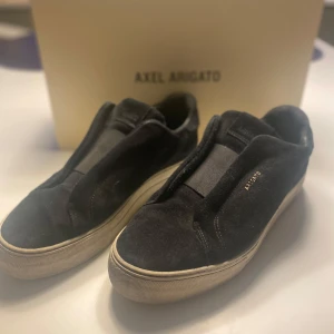 Svarta sneakers från Axel Arigato - Stilrena svarta sneakers från Axel Arigato i mocka med vit platt sula. Skorna har minimalistisk design utan snörning och diskret guldtext på sidan. Perfekta för dig som gillar en clean look och vill ha något som funkar till det mesta. 