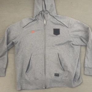 Grå hoodie från Nike med dragkedja - Snygg grå hoodie från Nike med hel dragkedja och huva med snörning. Tröjan har orange Nike-logga på bröstet och en svart patch med ett Lejon på. Mjuk insida och två fickor framtill. Perfekt för chill dagar eller när du vill ha en sportig look. Storlek XXL men passar XL också. Pris kan diskuteras! 