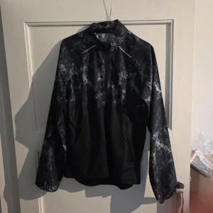 Svart vindjacka från SOC, stl S - Svart vindjacka från SOC med grått mönster och reflekterande detaljer. Jackan har hel dragkedja framtill, hög krage och är tillverkad i lätt syntetmaterial som andas. Perfekt för löpning eller utomhusaktiviteter. Snygg och sportig look med lång ärm.