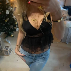 Svart volangig blus med v-ringning - Säljer en svart blus med djup v-ringning och volanger i transparent tyg. Toppen är ärmlös och har en feminin, flowy look som passar perfekt till jeans eller kjol. Snygg och trendig modell med lätt croppad passform.