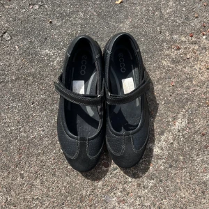 Svarta ballerina med kardborre - Snygga svarta ballerina från ECCO med kardborreband över foten. Skorna har rund tå, perforerade detaljer på sidorna och kontrastsömmar. Tillverkade i skinn och syntet, med mjuk och flexibel sula som ger bra komfort hela dagen. Sparsamt använda, nypris 800:-