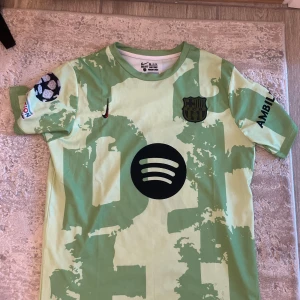 Barcelona Lamine Yamal Champions League tröja - Säljer en Barcelona Champions League fotbollströja med Lamine Yamal och nummer 10 på ryggen. Tröjan är ljusgrön med mörkgröna camouflage-detaljer, svarta tryck och flera loggor, bland annat Spotify och UEFA Foundation. Kortärmad och tillverkad i lätt polyester.