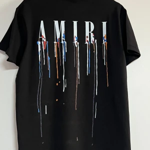 Svart AMIRI t-shirt med färgstänk - Svart t-shirt från AMIRI med stort tryck på ryggen och mindre tryck på bröstet. Trycket består av AMIRI-loggan och färgstänk i blått, vitt, orange och rött som rinner ner. Rund hals och korta ärmar, tillverkad i mjuk bomull. Perfekt för dig som gillar streetwear med edge.