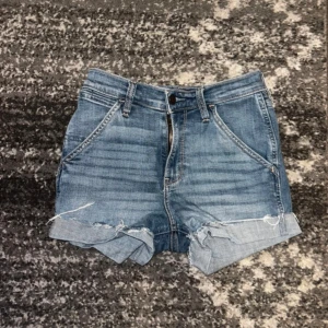 Blå jeansshorts Hollister - Snygga blå jeansshorts från Hollister med slitna detaljer och uppvikta benslut. Klassisk femficksmodell med dragkedja och knapp framtill. Perfekta för varma dagar och en avslappnad stil.