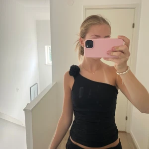 Svart oneshoulder topp från Amisu strl Xs - Svart oneshoulder topp från Amisu med en snygg volangblomma på axeln. Toppens material är stretchigt och figurnära. Passformen är tight och axelbandslös på ena sidan för en cool look. Super fin💗 strl Xs