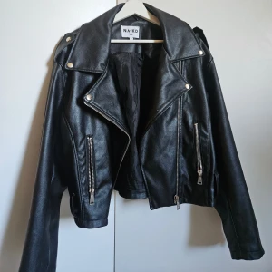 Svart skinnjacka från NA-KD, stl 36 - Säljer en svart skinnjacka från NA-KD i storlek 36. Jackan har klassisk bikerstil med silvriga dragkedjor, bred krage och coola detaljer vid axlarna. Perfekt för att lyfta vilken outfit som helst.