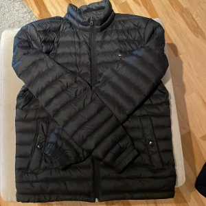 Svart pufferjacka Tommy Hilfiger S - Svart pufferjacka från Tommy Hilfiger i storlek S. Jackan har quiltad design, dragkedja framtill och två fickor med knappstängning. Klassisk ståkrage och diskret logga på bröstet. Perfekt för kyliga dagar när du vill hålla stilen.