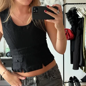 Svart linnetopp med fickdetalj - Snygg svart linnetopp med ärmlös design och rund halsringning. Toppen har en unik skärning nertill och en liten fickdetalj framtill. 